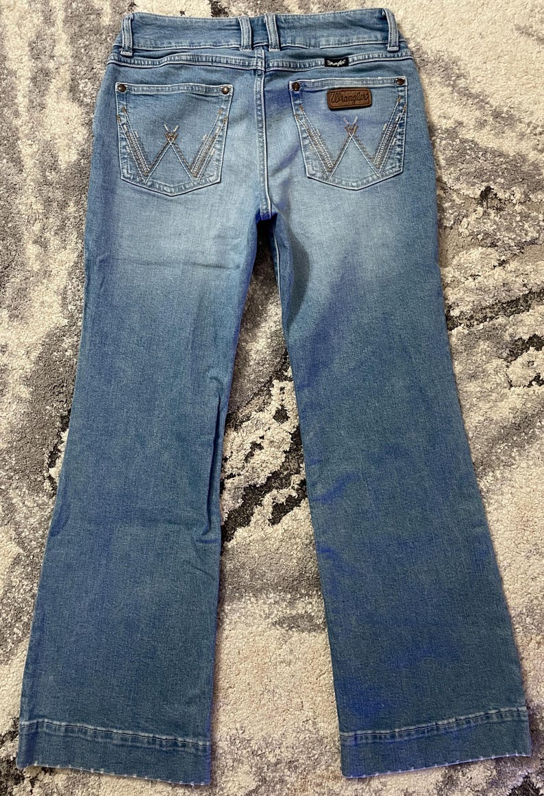 Puede incluir: Vaqueros de mezclilla azul claro con pernera acampanada. Los bolsillos traseros presentan un dise&ntilde;o de "W" cosido. Un parche de cuero marr&oacute;n con el logotipo "Wrangler" es visible en la cintura trasera.
