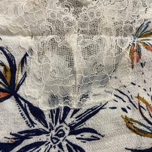 Puede incluir: Primer plano de una tela blanca con un estampado floral y ribete de encaje. La tela presenta grandes flores azul marino con detalles intrincados y acentos de naranja y amarillo. El ribete de encaje es delicado y a&ntilde;ade un toque de elegancia.