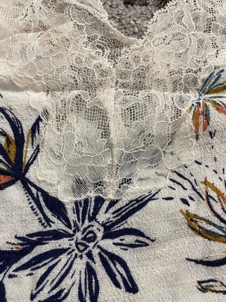 Puede incluir: Primer plano de una tela blanca con un estampado floral y ribete de encaje. La tela presenta grandes flores azul marino con detalles intrincados y acentos de naranja y amarillo. El ribete de encaje es delicado y a&ntilde;ade un toque de elegancia.