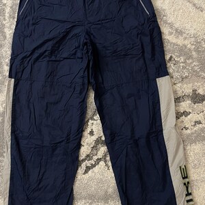 Pu&ograve; includere: Pantaloni sportivi blu navy con una striscia grigio chiaro sul lato. Il logo Nike &egrave; stampato verticalmente in verde scuro sulla parte inferiore della gamba. I pantaloni hanno tasche con cerniera e vita elasticizzata.