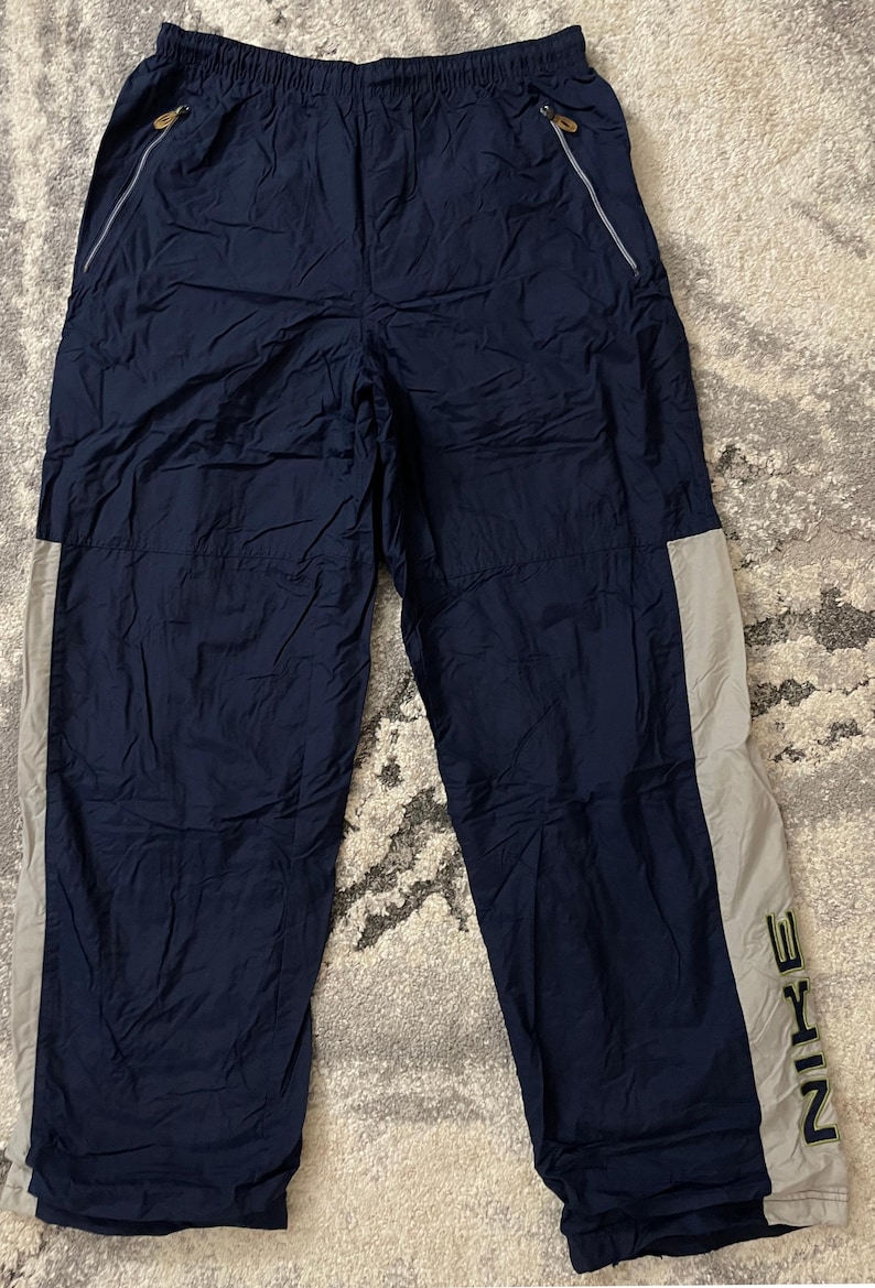 Pu&ograve; includere: Pantaloni sportivi blu navy con una striscia grigio chiaro sul lato. Il logo Nike &egrave; stampato verticalmente in verde scuro sulla parte inferiore della gamba. I pantaloni hanno tasche con cerniera e vita elasticizzata.