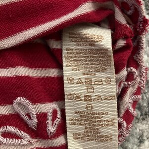 May include: Close-up of a red and white striped fabric with a tag. The fabric has white embroidered details along the edges. The tag displays care instructions and fabric composition.