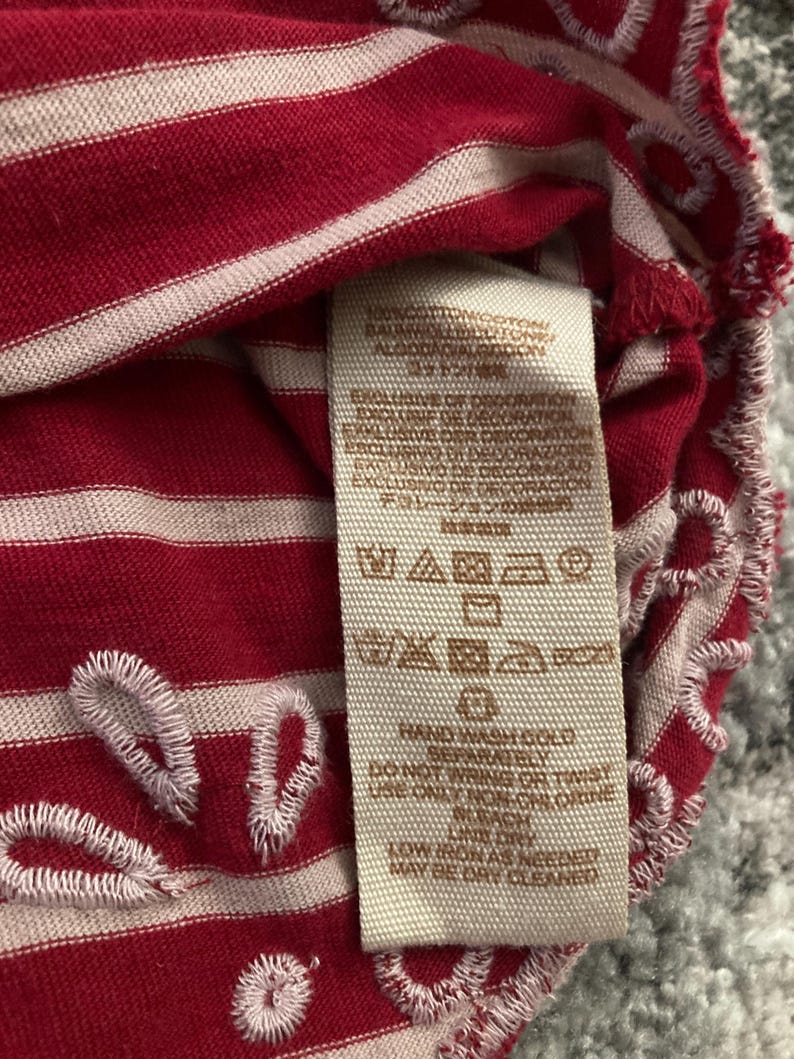 May include: Close-up of a red and white striped fabric with a tag. The fabric has white embroidered details along the edges. The tag displays care instructions and fabric composition.