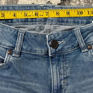 Puede incluir: Primer plano de unos vaqueros de mezclilla azul claro con una cinta m&eacute;trica amarilla en la cintura. La cinta mide aproximadamente 33 cm. Los vaqueros tienen cierre de bot&oacute;n y bolsillos.