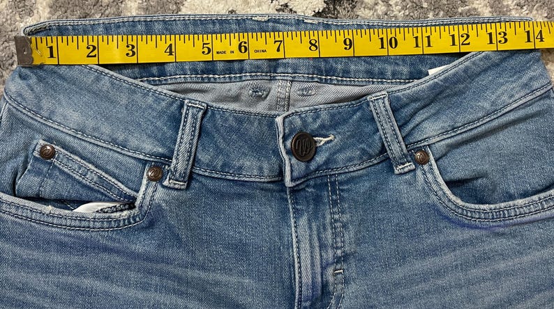 Puede incluir: Primer plano de unos vaqueros de mezclilla azul claro con una cinta m&eacute;trica amarilla en la cintura. La cinta mide aproximadamente 33 cm. Los vaqueros tienen cierre de bot&oacute;n y bolsillos.