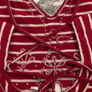 May include: Red and white striped top with a crisscross back design. The top features white embroidered floral patterns and a V-neckline. The fabric has a soft, textured appearance.