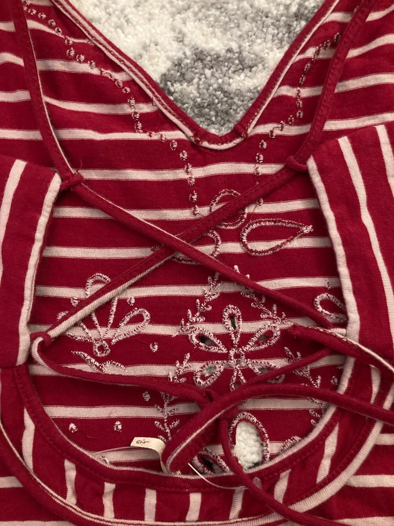 May include: Red and white striped top with a crisscross back design. The top features white embroidered floral patterns and a V-neckline. The fabric has a soft, textured appearance.