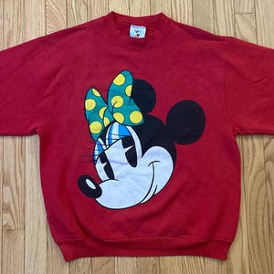 May include: A red sweatshirt featuring a large graphic of Minnie Mouse. The graphic shows Minnie's face with a black head, white face, and a bow with yellow polka dots on a green background. The sweatshirt has long sleeves.