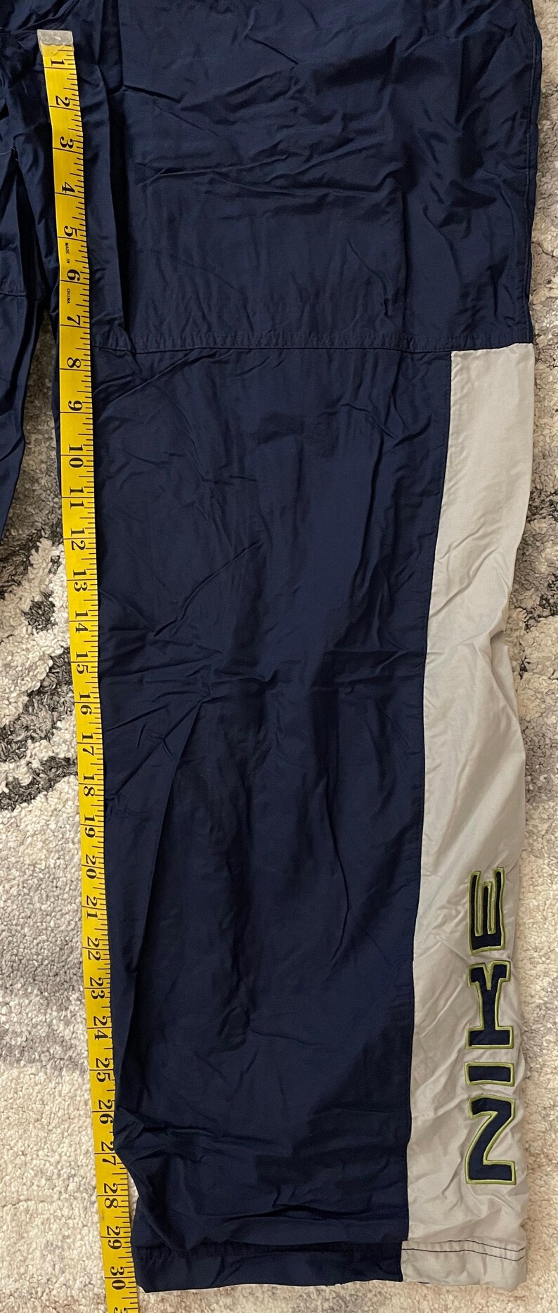 Pu&ograve; includere: Pantaloni sportivi blu navy e grigi con il logo Nike in verde. I pantaloni sono lunghi circa 76 cm, misurati con un metro a nastro giallo.