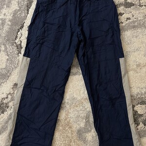 Pu&ograve; includere: Pantaloni sportivi blu navy con una striscia grigio chiaro sul lato. I pantaloni hanno una vita con coulisse e un piccolo logo dorato sulla parte superiore della gamba sinistra. I pantaloni sono realizzati in un materiale leggero, possibilmente nylon.