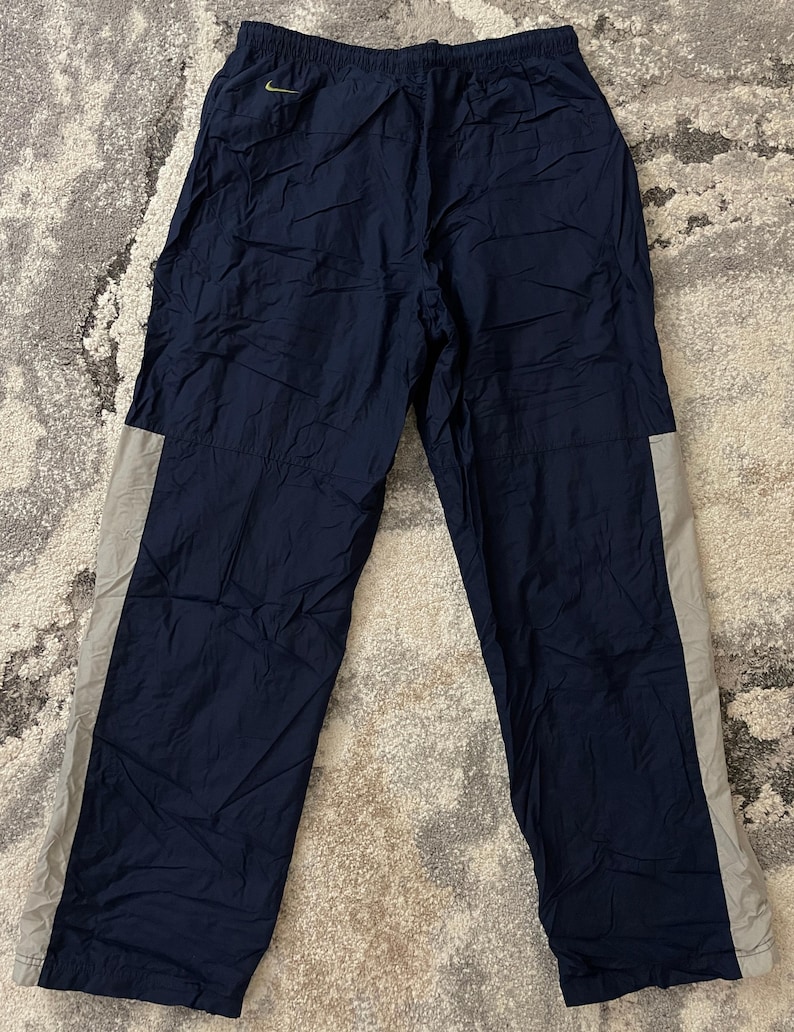Pu&ograve; includere: Pantaloni sportivi blu navy con una striscia grigio chiaro sul lato. I pantaloni hanno una vita con coulisse e un piccolo logo dorato sulla parte superiore della gamba sinistra. I pantaloni sono realizzati in un materiale leggero, possibilmente nylon.