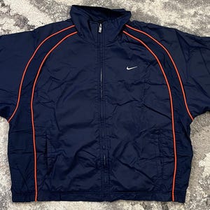 May include: Navy blue Nike track jacket with orange trim. The jacket has a full-length zipper, a small white Nike logo on the chest, and a high collar. The jacket's sleeves are long and the jacket is spread out on a patterned surface.