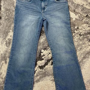 Puede incluir: Vaqueros de mezclilla azul claro con pernera acampanada. Los vaqueros tienen cierre de bot&oacute;n y cremallera, bolsillos delanteros y traseros, y la marca "Wrangler" es visible en la cintura.