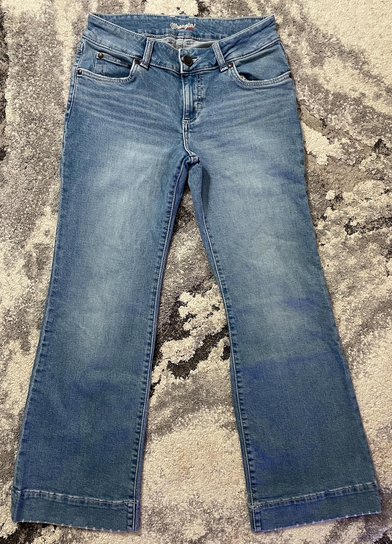 Puede incluir: Vaqueros de mezclilla azul claro con pernera acampanada. Los vaqueros tienen cierre de bot&oacute;n y cremallera, bolsillos delanteros y traseros, y la marca "Wrangler" es visible en la cintura.