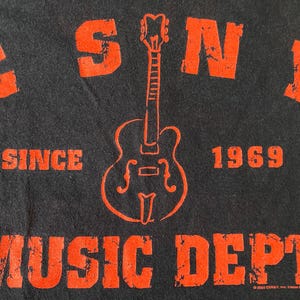 Könnte beinhalten: Schwarzes T-Shirt mit verblichenem orangefarbenem Text und einer Gitarrengrafik. Der Text lautet "CSNY MUSIC DEPT. SINCE 1969". Die Gitarre ist eine stilisierte Umrisszeichnung. Das Shirt hat eine Vintage-Retro-Ästhetik.