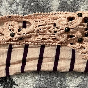 May include: Close-up of a long-sleeved shirt sleeve. The sleeve is striped with alternating light pink and dark purple stripes. The cuff features a decorative, light pink, openwork lace design with small, dark brown beads.