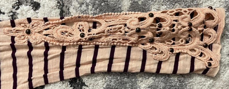 May include: Close-up of a long-sleeved shirt sleeve. The sleeve is striped with alternating light pink and dark purple stripes. The cuff features a decorative, light pink, openwork lace design with small, dark brown beads.