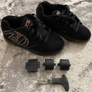 May include: Black Heelys roller skate shoes with brown soles and copper-colored accents. Includes a set of black wheels, a wheel removal tool, and a small black plate. The shoes have laces and the Heelys logo.