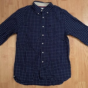 May include: A long-sleeve button-down shirt in a dark navy blue fabric featuring a white grid pattern. The shirt includes a pointed collar with button-down points, a front button placket, and long sleeves with buttoned cuffs, presenting a classic design.