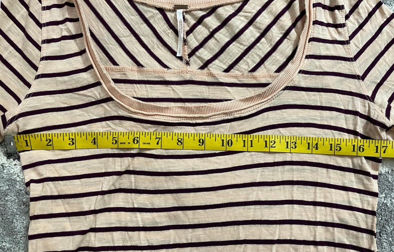 May include: A cream-colored, scoop-neck shirt with dark burgundy horizontal stripes. The shirt is measured with a yellow tape measure, showing a width of approximately 17 inches.