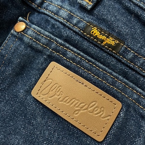 May include: Close-up of blue denim jeans with visible stitching and a brown leather patch embossed with the brand name "Wrangler." A black label with the "Wrangler" logo in gold is also visible.
