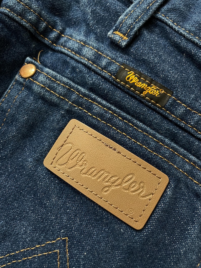 May include: Close-up of blue denim jeans with visible stitching and a brown leather patch embossed with the brand name "Wrangler." A black label with the "Wrangler" logo in gold is also visible.