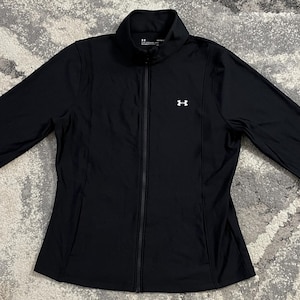 May include: A black long-sleeve athletic jacket with a full-length front zipper, a high collar, and a small white Under Armour logo on the upper left chest. The jacket features fitted sleeves with thumbhole cuffs and a smooth, solid black fabric.