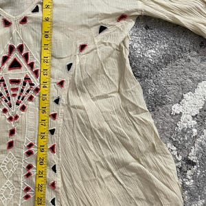 Może przedstawiać: Kremowa tunika z dekoltem w serek i długimi rękawami. Tunika posiada haftowane geometryczne wzory w kolorze czarnym, czerwonym i białym. Długość tuniki to około 76 cm.