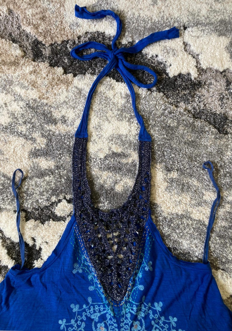K&ouml;nnte beinhalten: Ein blaues Neckholder-Top mit geh&auml;keltem Ausschnitt und Kordelverschluss. Das Top hat ein detailliertes H&auml;keldesign in einem dunkleren Blauton mit einem helleren blauen Stickmuster auf der Vorderseite.