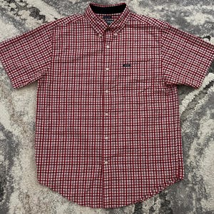 May include: Red and white checkered short-sleeve button-down shirt. The shirt has a button-down collar, a chest pocket, and the brand name "Chaps" embroidered on the pocket. The shirt is made of a lightweight fabric.