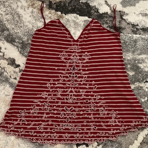 May include: A burgundy and white striped camisole with a floral design. The camisole features thin spaghetti straps and a scalloped hemline. The floral design is embroidered in white and is centered on the front of the camisole.