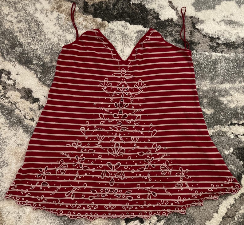 May include: A burgundy and white striped camisole with a floral design. The camisole features thin spaghetti straps and a scalloped hemline. The floral design is embroidered in white and is centered on the front of the camisole.