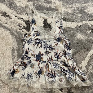 Puede incluir: Camiseta sin mangas blanca con estampado floral con ribete y espalda de encaje. La camiseta sin mangas presenta un escote en V y un estampado floral en azul marino, marr&oacute;n y naranja. El ribete de encaje es blanco.
