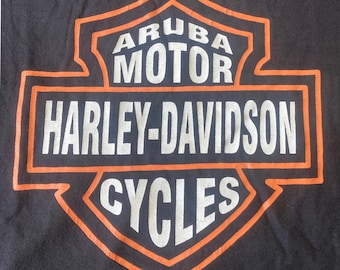 May include: Black t-shirt featuring an orange and white Harley-Davidson Aruba Motor Cycles logo. The logo is a shield shape with the words "ARUBA MOTOR" at the top, "HARLEY-DAVIDSON" in the middle, and "CYCLES" at the bottom.