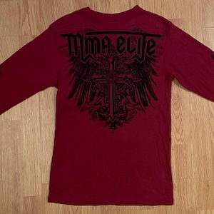 Może przedstawiać: Ciemnoczerwona koszulka z długim rękawem z czarnym nadrukiem graficznym na klatce piersiowej, zawierającym napis MMA ELITE nad stylizowanym wzorem krzyża i skrzydeł. Dodatkowe czarne grafiki krzyży znajdują się na zewnętrznej stronie obu rękawów.