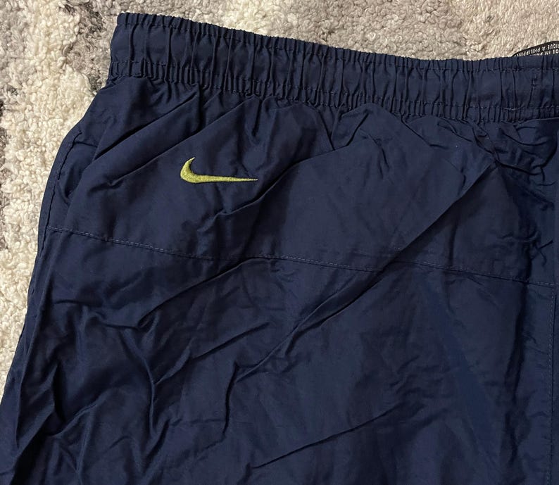Pu&ograve; includere: Pantaloni sportivi blu navy con elastico in vita e tasca laterale. Un logo Nike swoosh giallo &egrave; visibile sulla parte superiore sinistra dei pantaloni. I pantaloni sono realizzati in materiale leggero, possibilmente nylon.