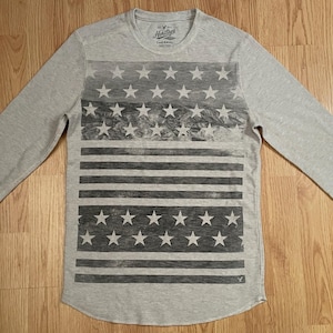 Könnte beinhalten: Ein hellgraues Langarm-Thermo-Shirt mit einem zentralen Grafikdruck eines Flaggendesigns mit weißen Sternen und dunkelgrauen horizontalen Streifen. Das Shirt hat einen Rundhalsausschnitt, einen abgerundeten Saum und das Heritage-Markenetikett.