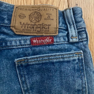 以下が含まれることがあります： 「Wrangler Authentic Jeans」と書かれた茶色の革パッチが付いた、青いデニムジーンズのクローズアップ。赤い「Wrangler」タグがバックポケットに縫い付けられています。ジーンズはクラシックな5ポケットデザインです。