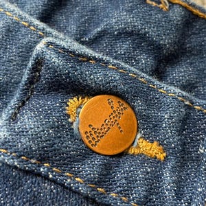 May include: Close-up of a blue denim fabric with a copper-colored button. The button is round with a textured design and is sewn onto the denim with yellow thread. The denim has visible stitching.