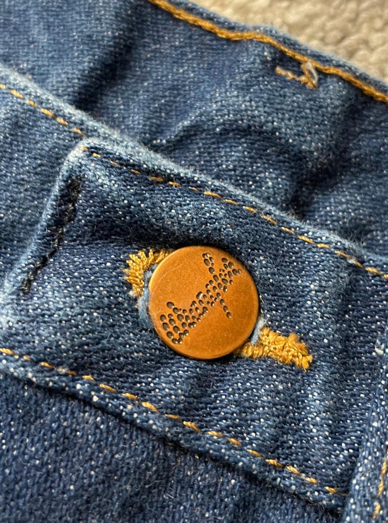 May include: Close-up of a blue denim fabric with a copper-colored button. The button is round with a textured design and is sewn onto the denim with yellow thread. The denim has visible stitching.