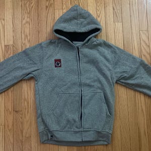 Könnte beinhalten: Ein grauer Kapuzenpullover mit Reißverschluss, einer schwarz gefütterten Kapuze und einem kleinen Aufnäher mit einem Smiley. Der Hoodie hat eine Vordertasche und ein angebrachtes Etikett. Das Kleidungsstück liegt auf einer Holzoberfläche.