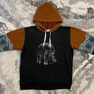 Peut inclure: Un sweat à capuche noir avec une capuche et le haut des manches marron, orné d'un motif de loups devant une pleine lune sur la poitrine. Les manches présentent une bande à motif géométrique bleu sarcelle, rouge et blanc, avec des cordons blancs.