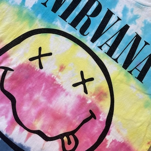May include: A tie-dye t-shirt featuring the word "NIRVANA" in large black letters. The shirt has a smiley face graphic with crossed-out eyes and a tongue sticking out. The background is a blend of blue, yellow, pink, and white.