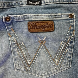 Puede incluir: Primer plano del bolsillo trasero de unos vaqueros azul claro. El bolsillo presenta un parche de cuero marr&oacute;n con el logotipo "Wrangler" y costuras decorativas. Los vaqueros tienen un dise&ntilde;o cl&aacute;sico de cinco bolsillos.
