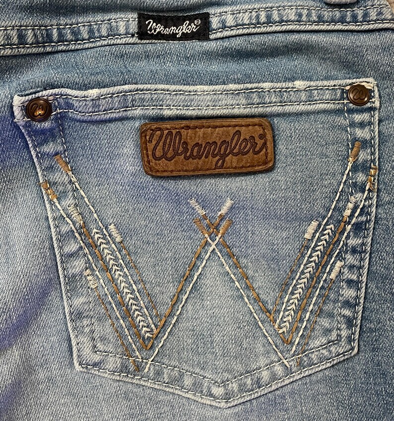 Puede incluir: Primer plano del bolsillo trasero de unos vaqueros azul claro. El bolsillo presenta un parche de cuero marr&oacute;n con el logotipo "Wrangler" y costuras decorativas. Los vaqueros tienen un dise&ntilde;o cl&aacute;sico de cinco bolsillos.