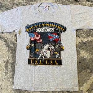 May include: A light gray short-sleeve t-shirt featuring a graphic of the Gettysburg Campaign with the text "GETTYSBURG JULY 1-3, 1863 CAMPAIGN" and "U.S.A. C.S.A." The illustration depicts two soldiers on horseback holding flags.