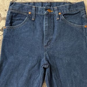 May include: Close-up of a pair of dark blue denim jeans. The jeans have classic five-pocket styling with copper-colored rivets and a button closure. The fabric appears to be a medium-weight denim.
