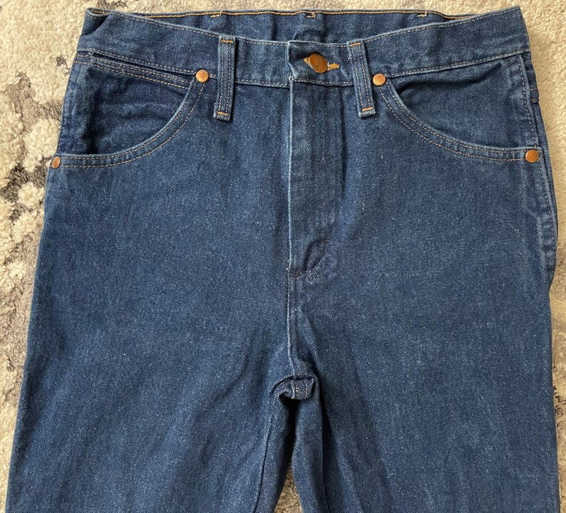 May include: Close-up of a pair of dark blue denim jeans. The jeans have classic five-pocket styling with copper-colored rivets and a button closure. The fabric appears to be a medium-weight denim.