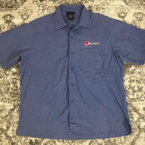 May include: A blue short-sleeved button-down shirt with a Pepsi logo embroidered on the left chest. The shirt has a pocket on the left side.