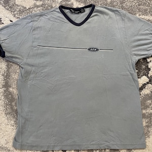 Puede incluir: Una camiseta gris claro de manga corta con cuello en V y ribetes azul marino oscuro en el cuello y las mangas. El pecho presenta un gráfico de línea horizontal con un logotipo ovalado que contiene el texto American Eagle y AE con una estrella.